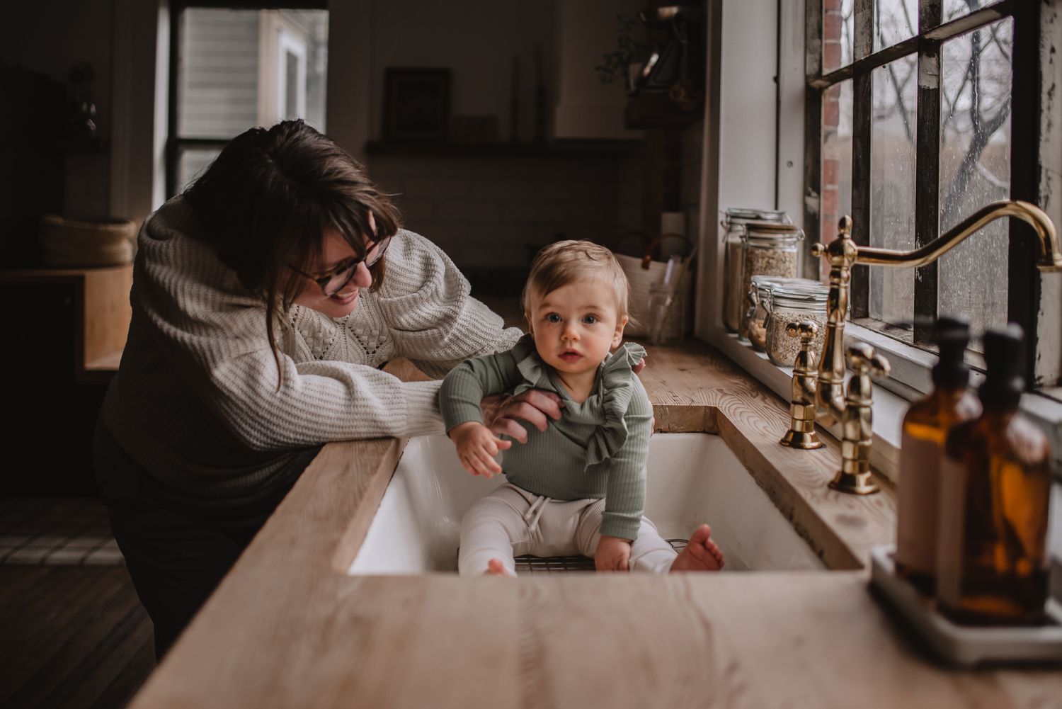 A tender kitchen scene with people by a farmhouse sink near a window.