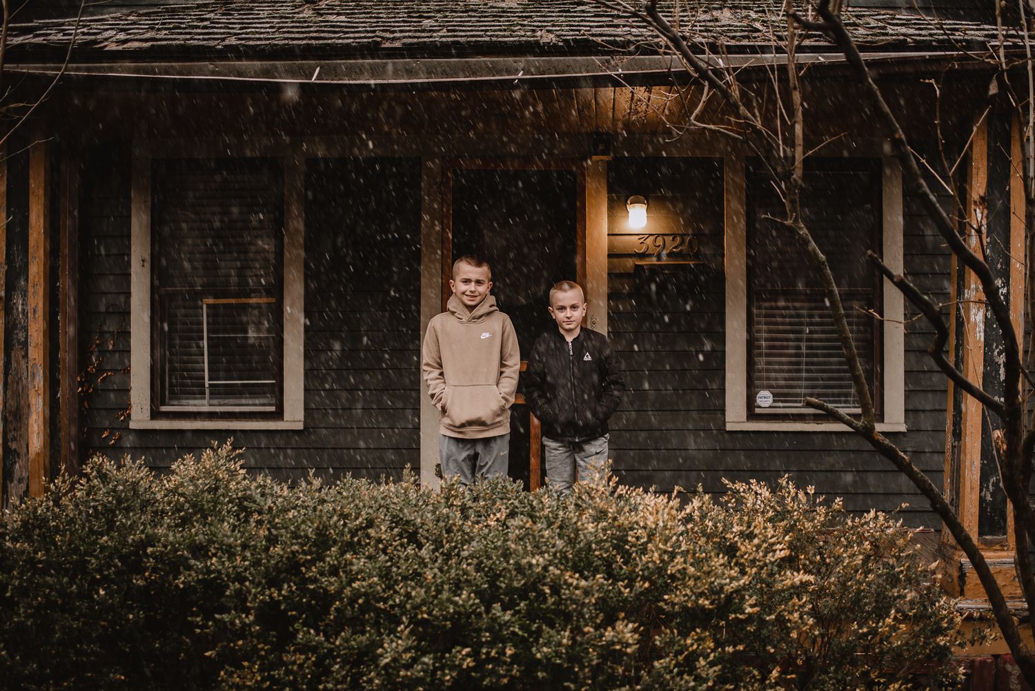 Two people standing in front of a rustic house exterior during snowfall.