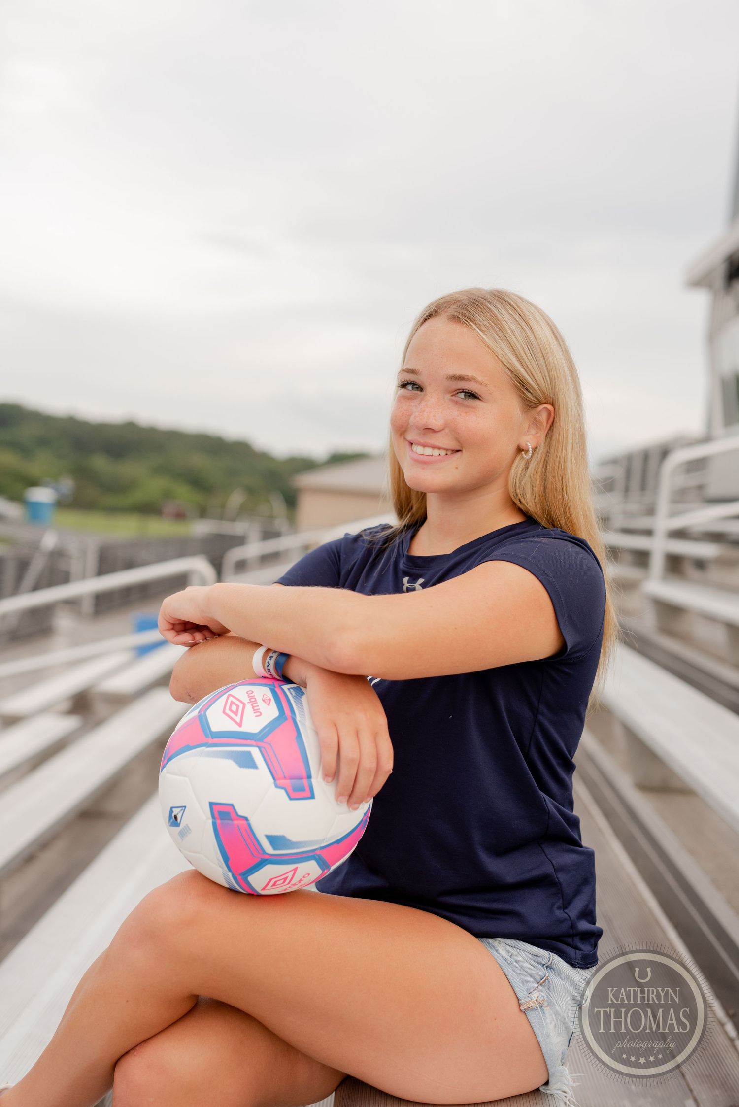 Athlete in navy jersey poses with soccer ball on stadium bleachers during sports portrait session.