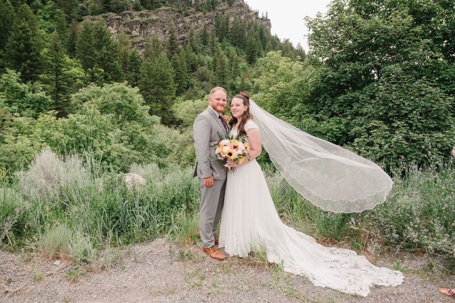Newlyweds pose on a mountain path as the bride's veil flows in the breeze.