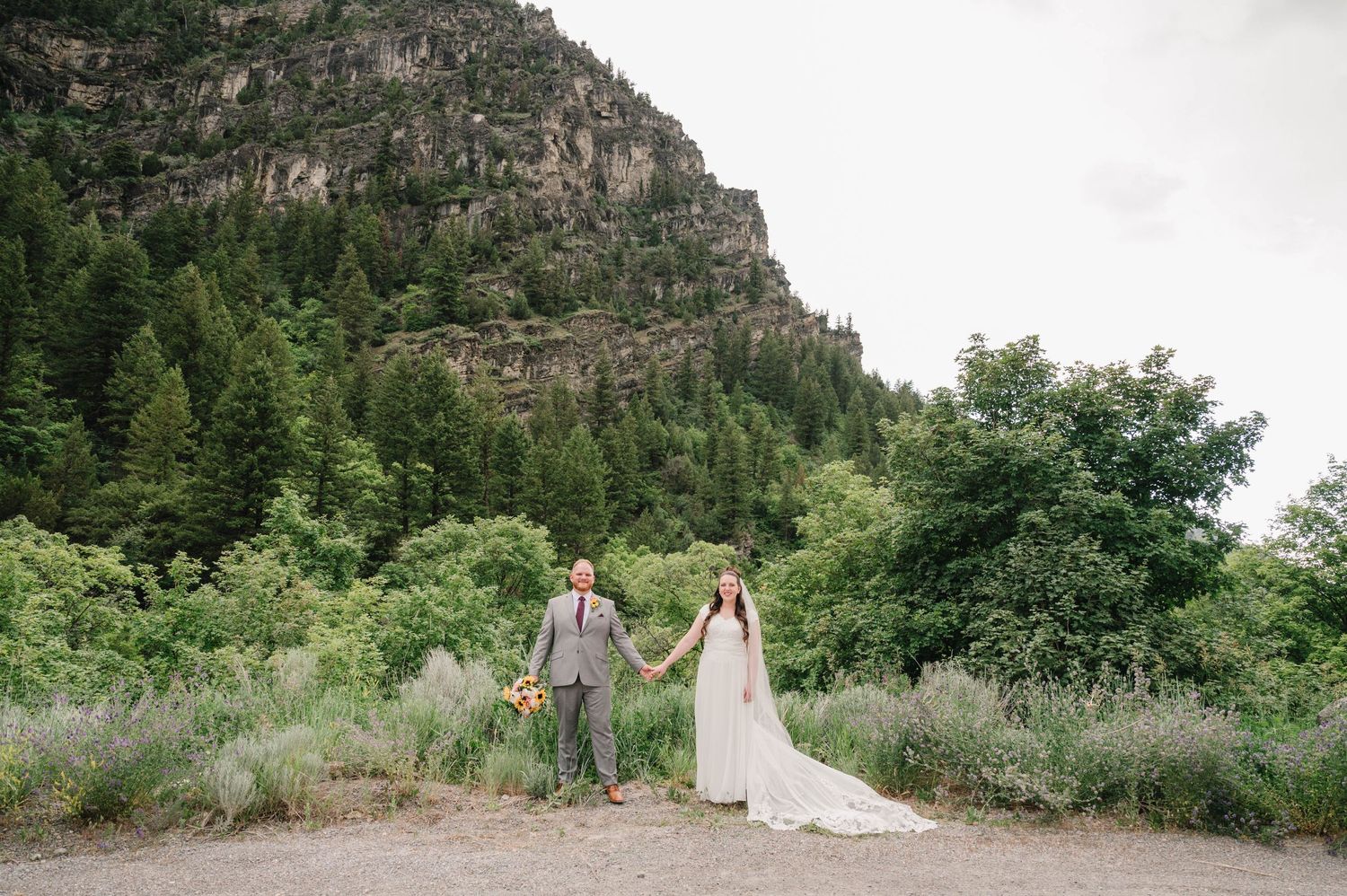 Couple holds hands while walking along a mountain trail in wedding attire.
