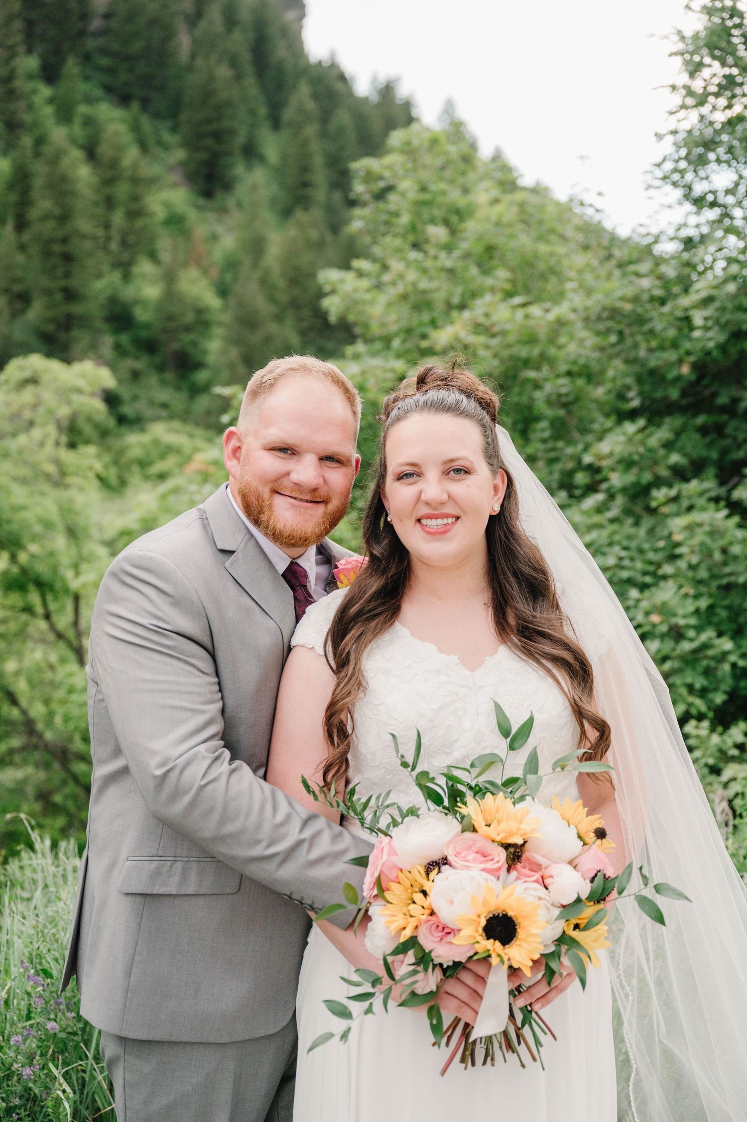 Wedding couple smiles while holding a vibrant bouquet featuring sunflowers against a mountain backdrop.