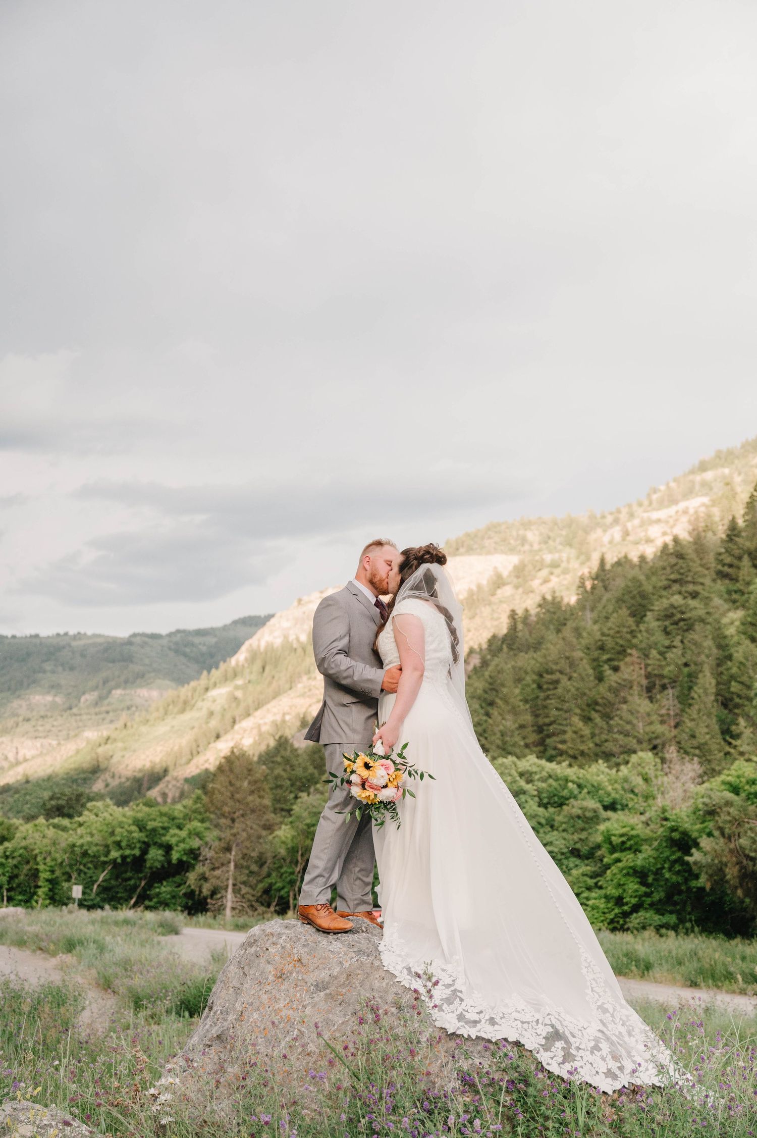 Newlyweds share an intimate moment while standing on a rock with mountain views.