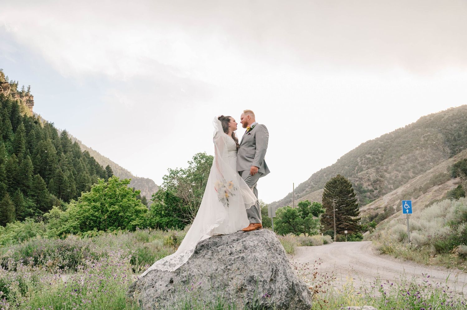 Bride and groom pose on a large boulder with mountainous landscape behind them.