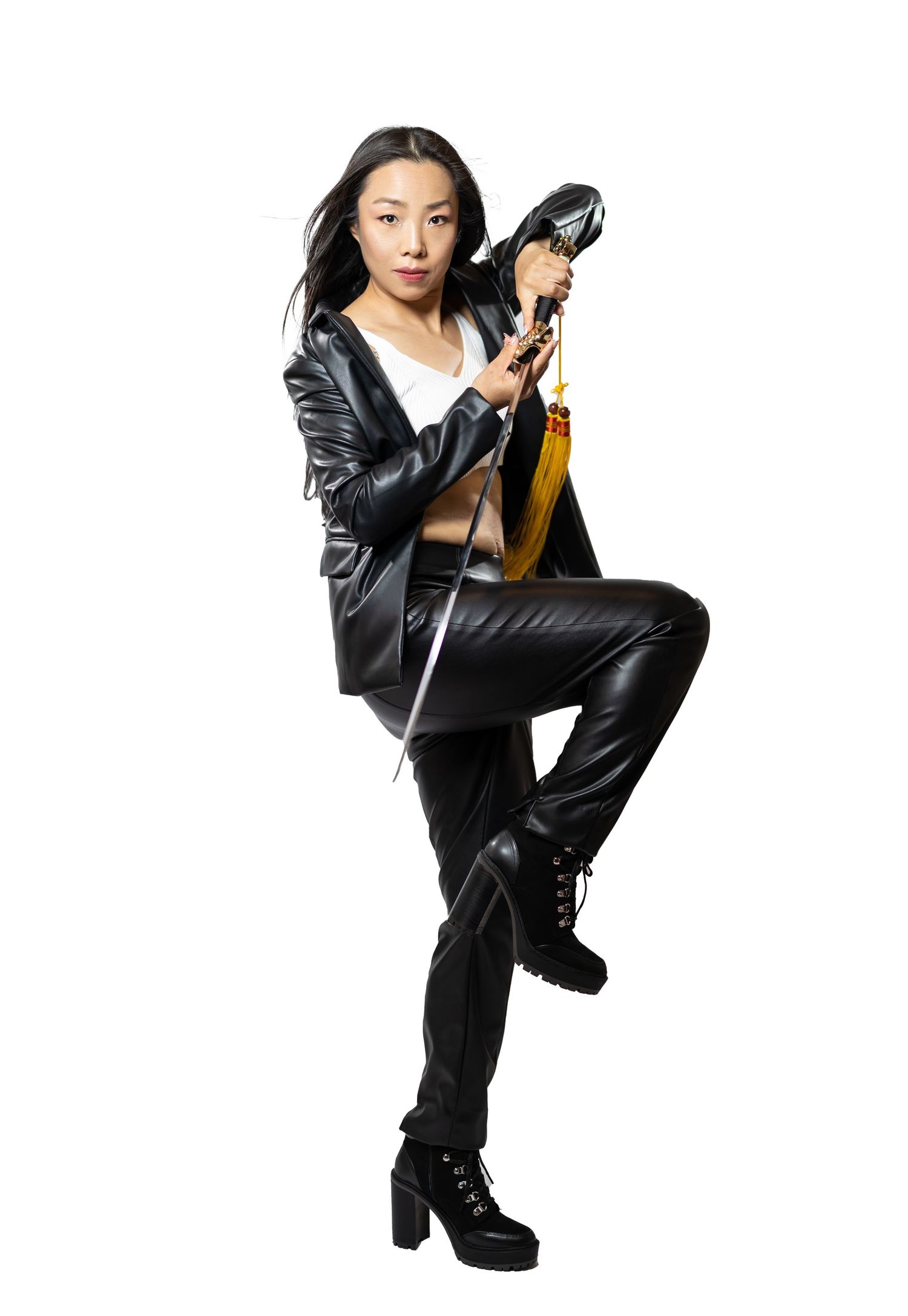 Stylish model poses with nunchucks wearing black leather outfit and boots against white background.