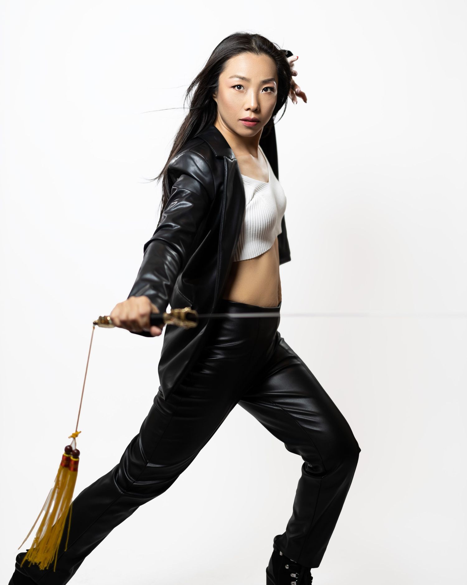 Athletic figure in black leather ensemble performs martial arts stance with nunchucks on white background.