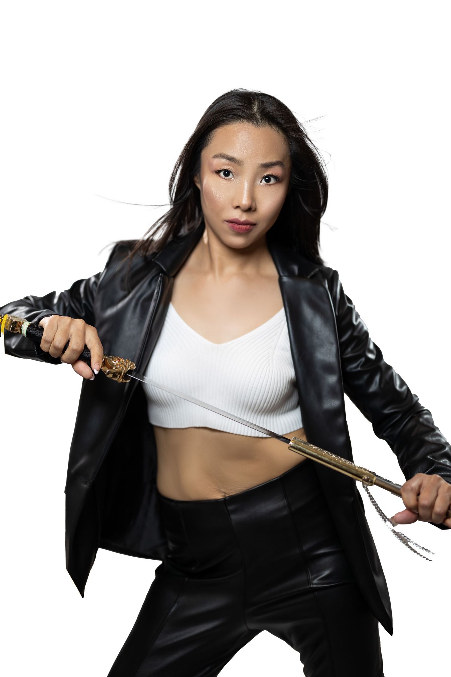 Fashion model in black leather outfit and white crop top strikes dynamic pose with nunchucks against white backdrop.