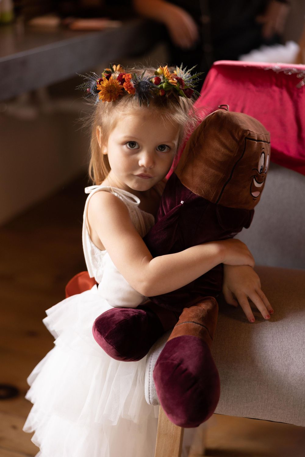 A young flower girl in white dress holding burgundy plush toy wears a fall floral crown.