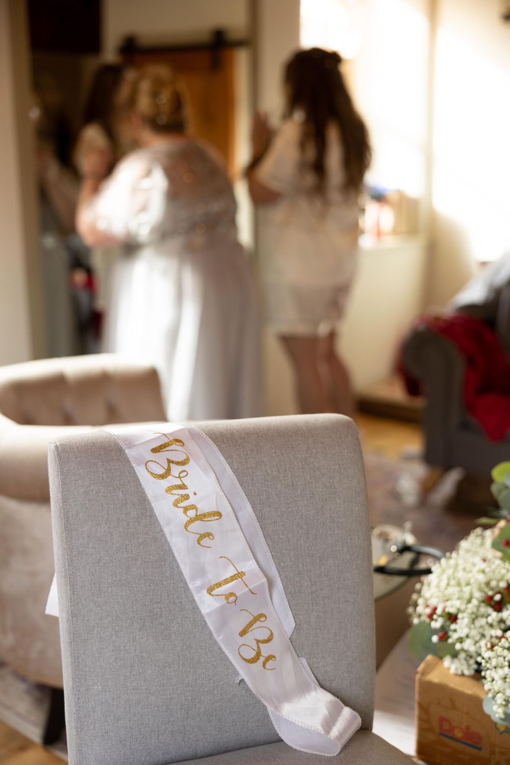 A white 'Bride to Be' sash rests on a light gray chair with wedding preparations happening in the background.