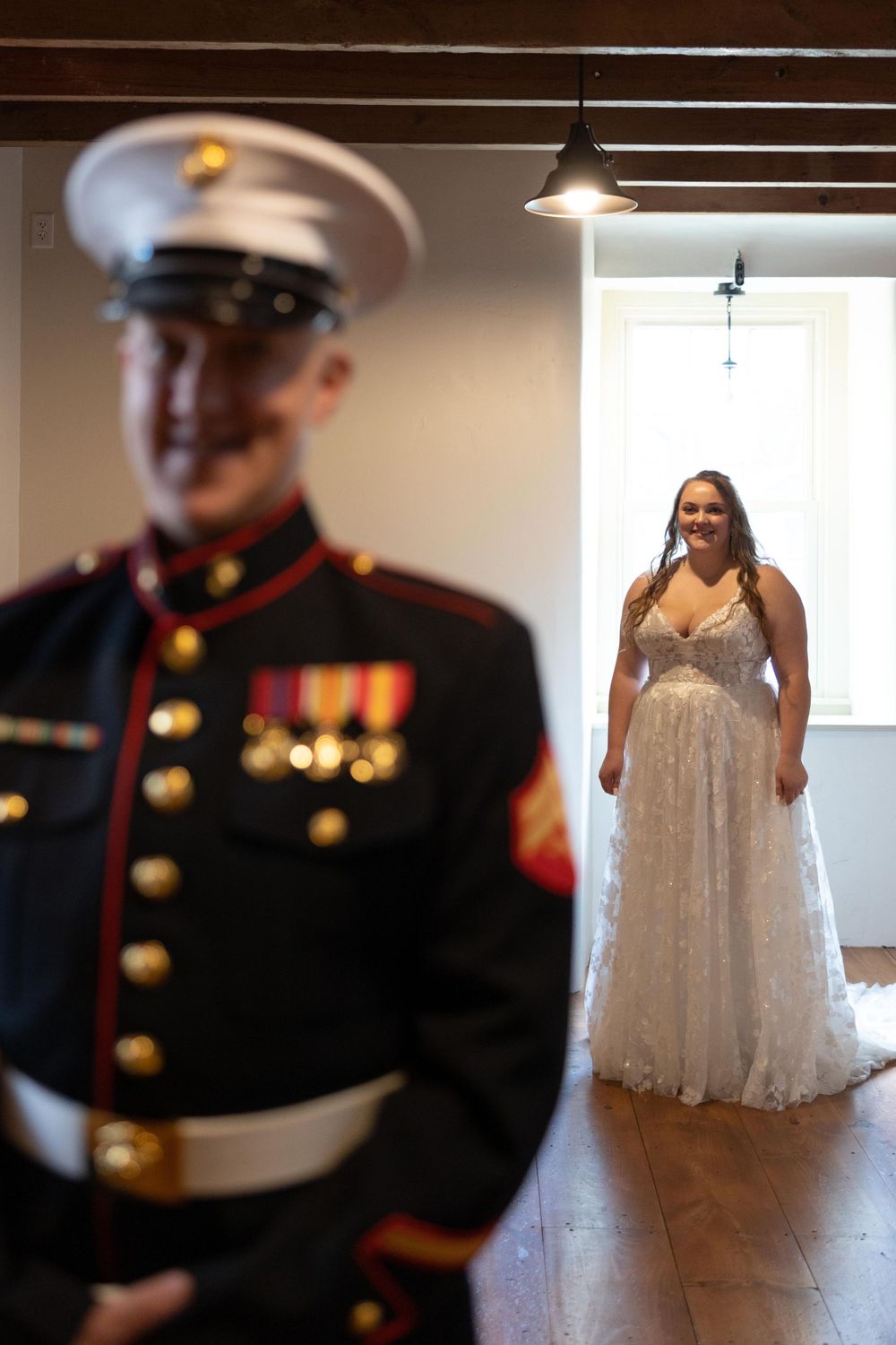 A military service member in formal dress uniform stands in focus while a bride waits in the background.