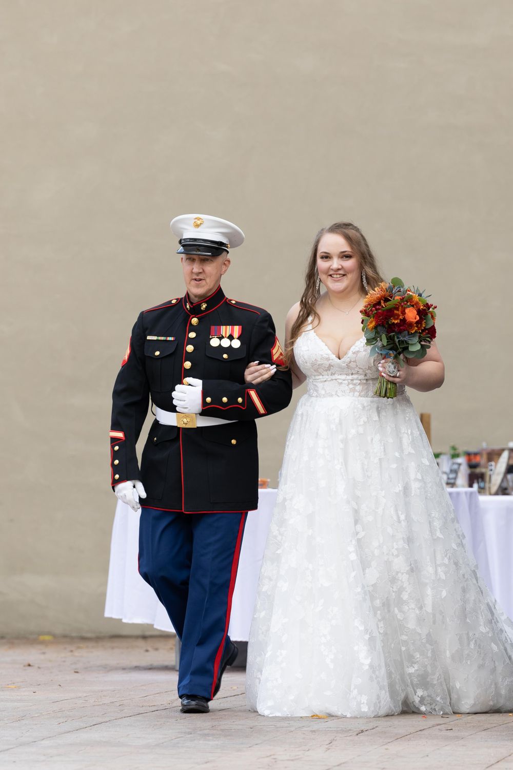 Marine officer in dress blues escorts bride in lace gown holding orange bouquet along beige wall.