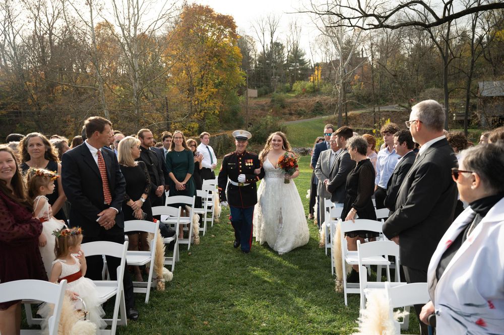 Outdoor autumn wedding ceremony with white chairs and guests on grass.