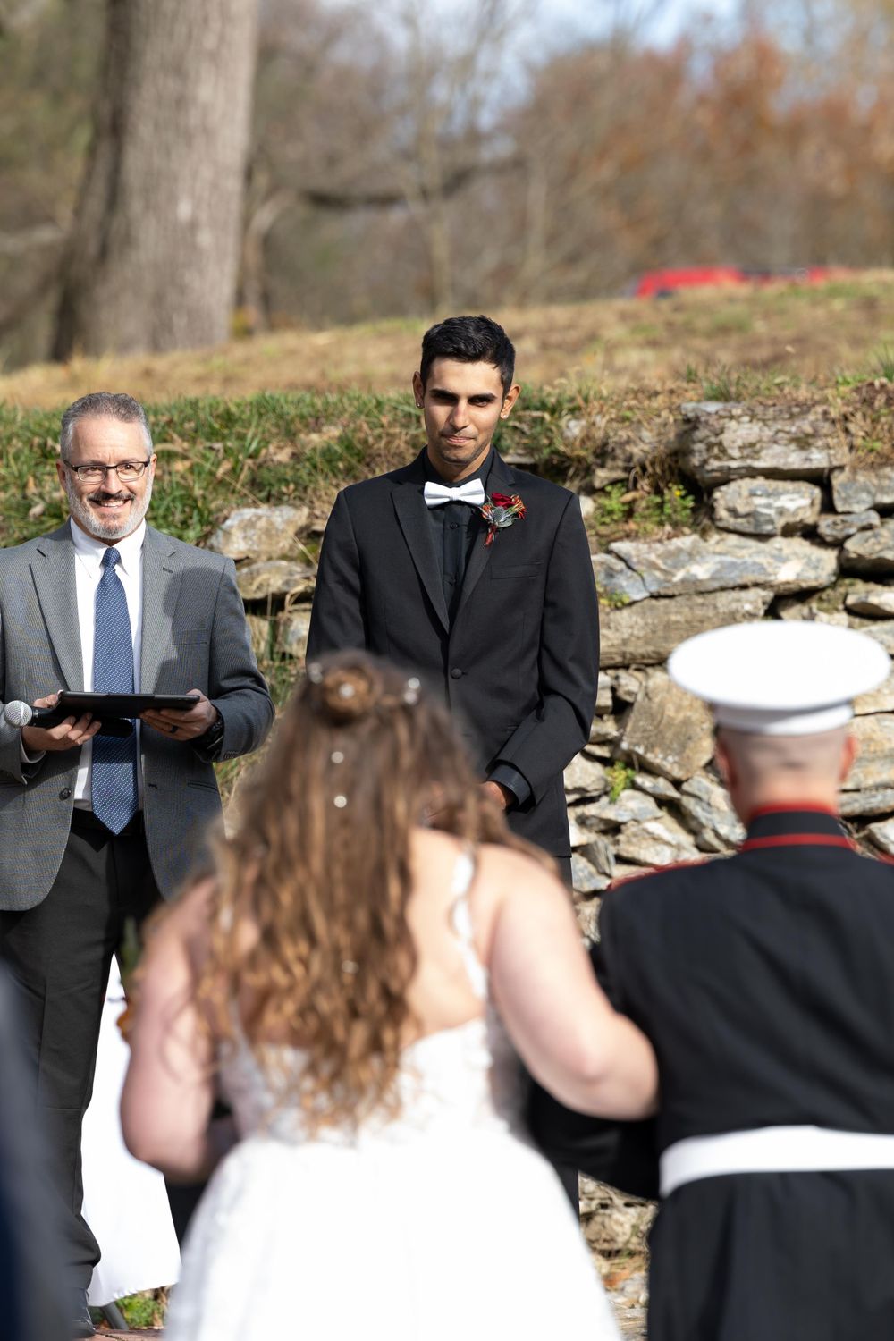 Outdoor wedding ceremony with officiant, groom, and Marine standing at stone wall altar.