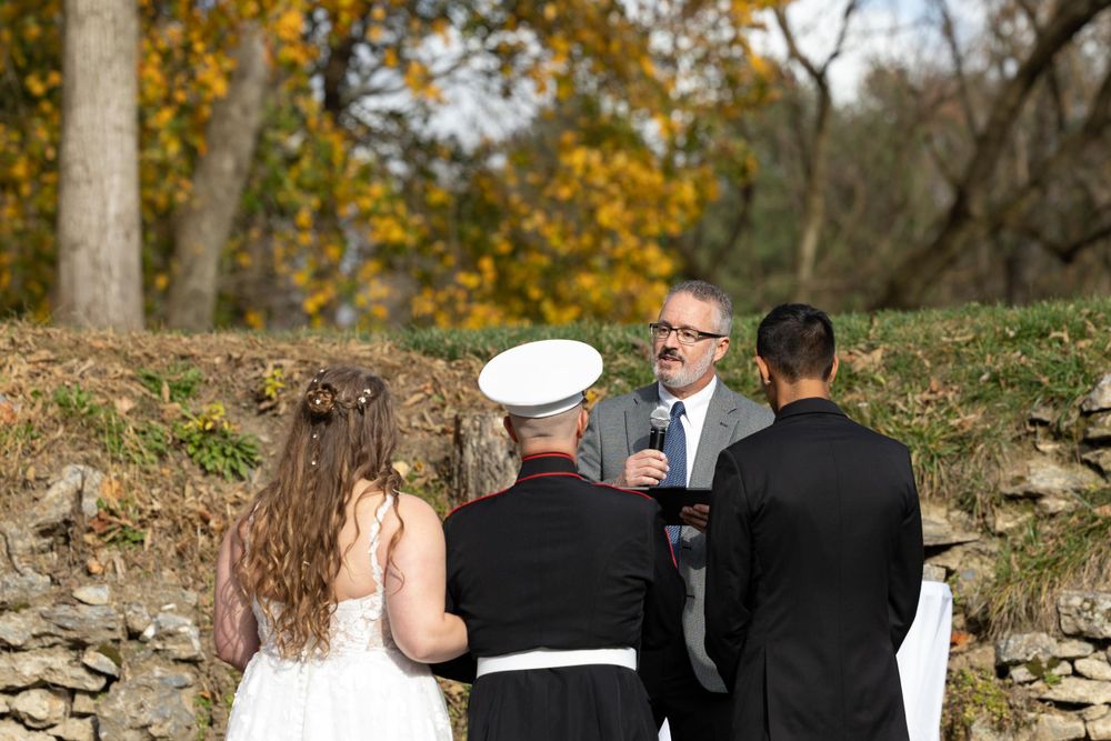 Intimate outdoor wedding ceremony with stone wall backdrop in fall foliage setting.