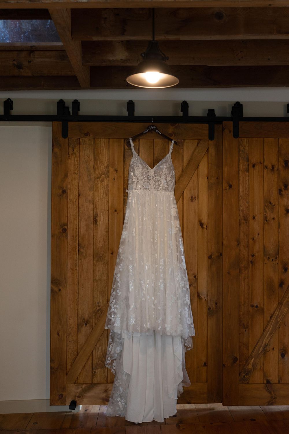 Lace wedding dress hanging on wooden barn door against white wall with pendant light.