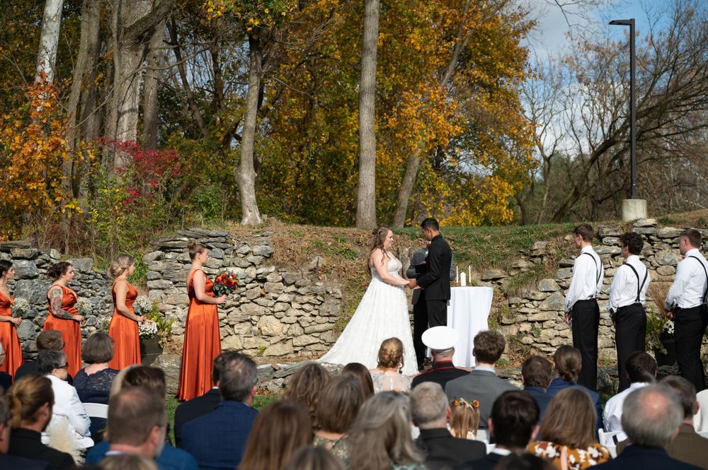 Outdoor wedding ceremony with stone wall backdrop and gathered guests during autumn season.