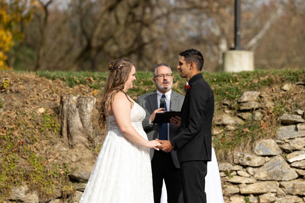 A wedding ceremony takes place outdoors on an autumn day with a stone wall backdrop.