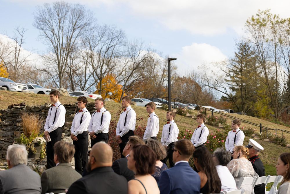 Wedding guests watch as groomsmen in white shirts and dark pants stand in a line during an outdoor ceremony.
