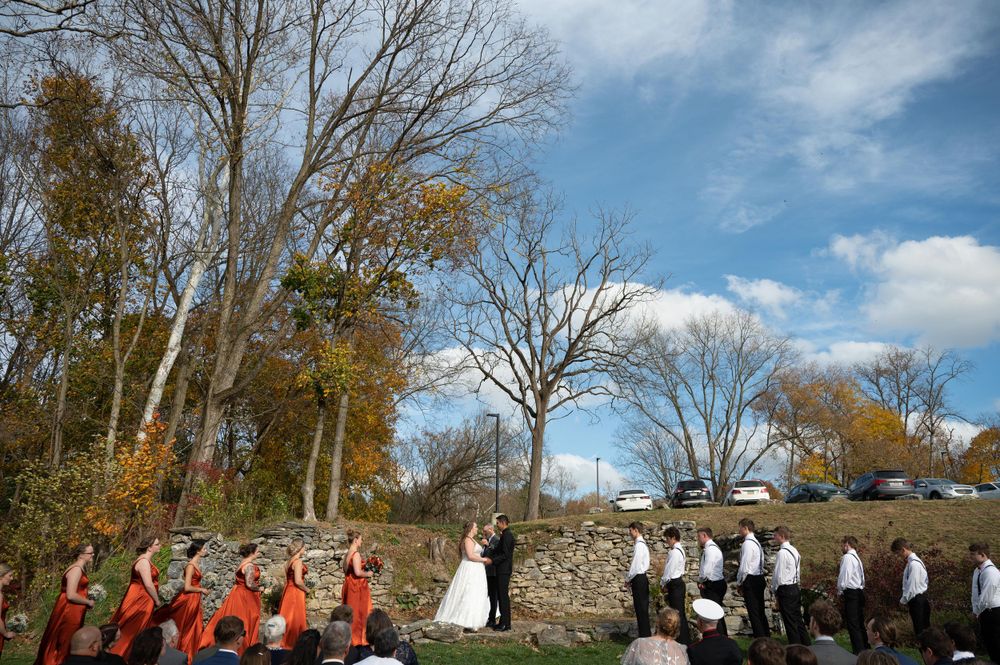 A wedding party stands in formation during an outdoor ceremony with fall foliage and blue skies.