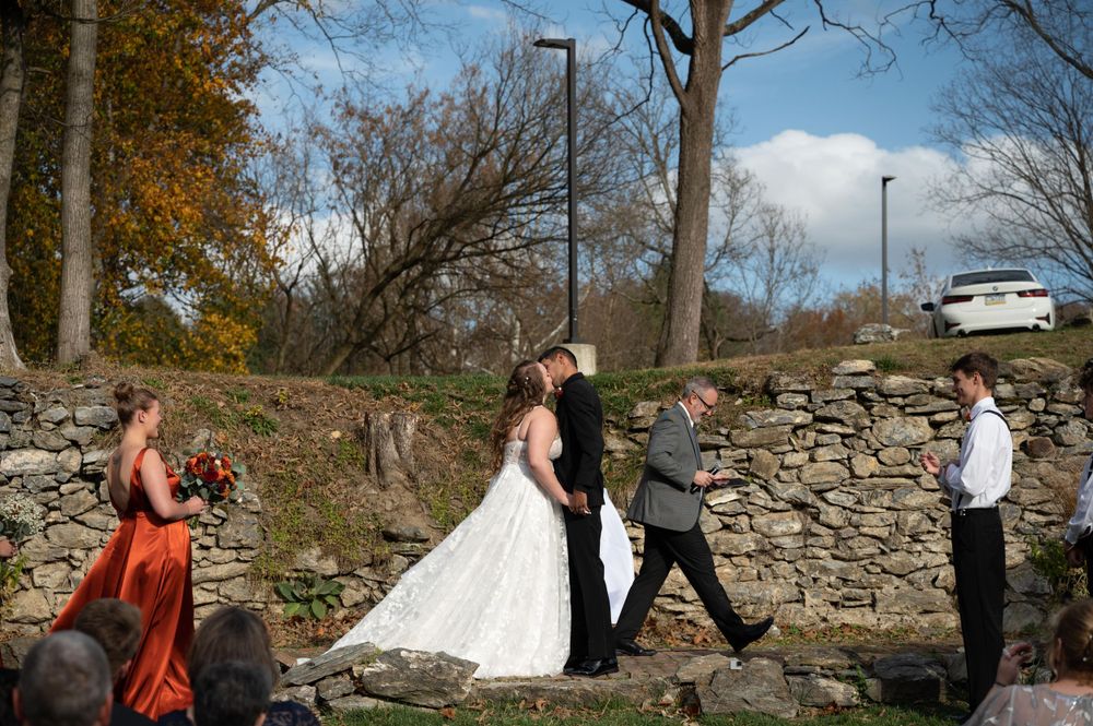 A wedding party processes across a grassy area with a stone wall and autumn trees in the background.