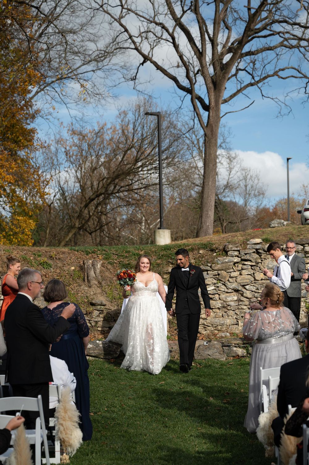 Newlyweds exit their outdoor ceremony while guests applaud against a backdrop of fall trees.