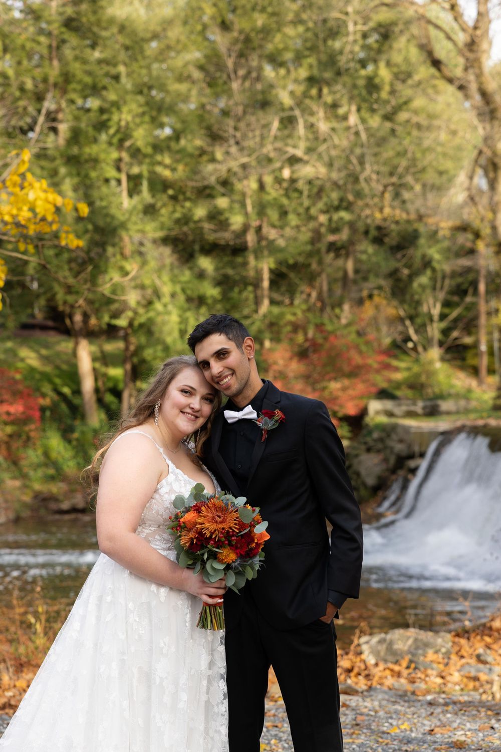 A couple poses for portraits by a waterfall surrounded by vibrant autumn foliage.