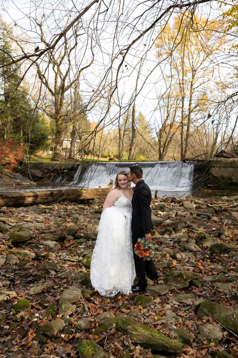 A wedding couple stands on rocky terrain with a serene waterfall and bare branches in the background.