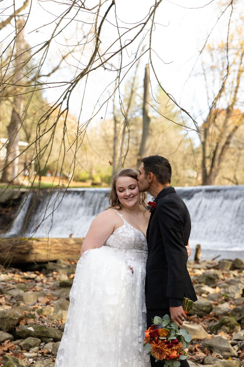 A romantic autumn wedding portrait with a cascading waterfall and bare tree branches overhead.