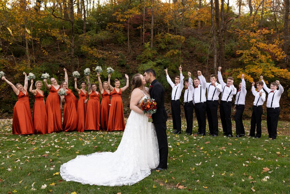 Wedding party in burgundy dresses and white shirts celebrates with raised arms on autumn grass.