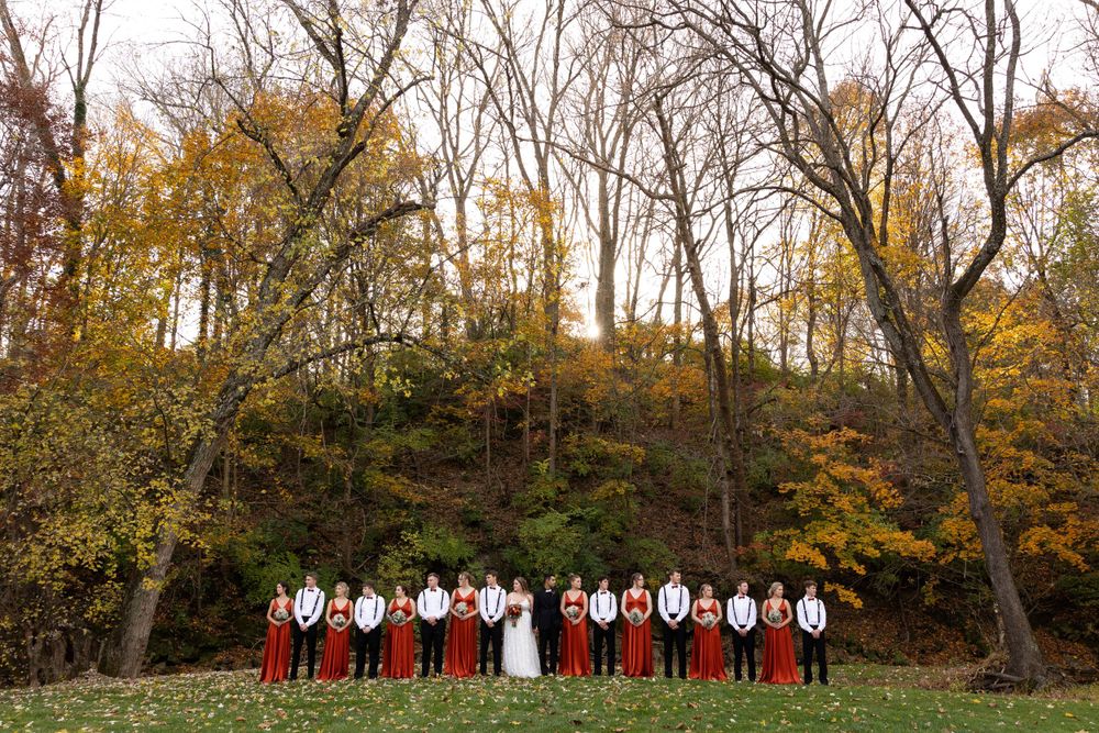 Wedding party lined up against autumn forest backdrop wearing red and white formal attire.