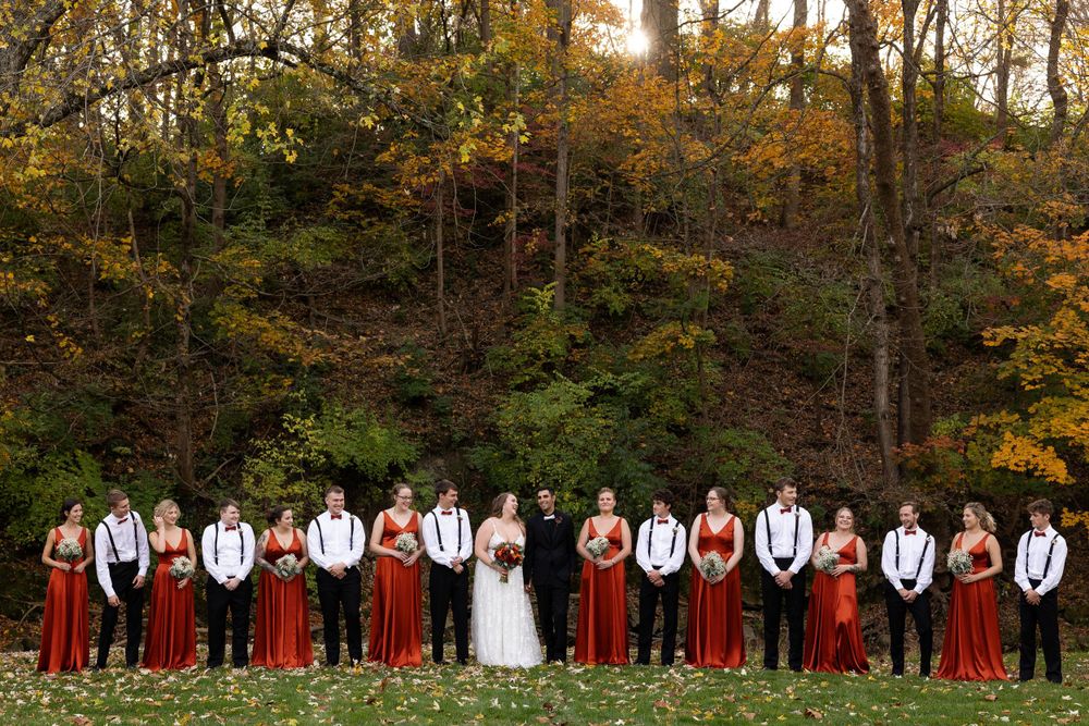 Full wedding party lined up in burgundy and white attire against autumn woodland backdrop.