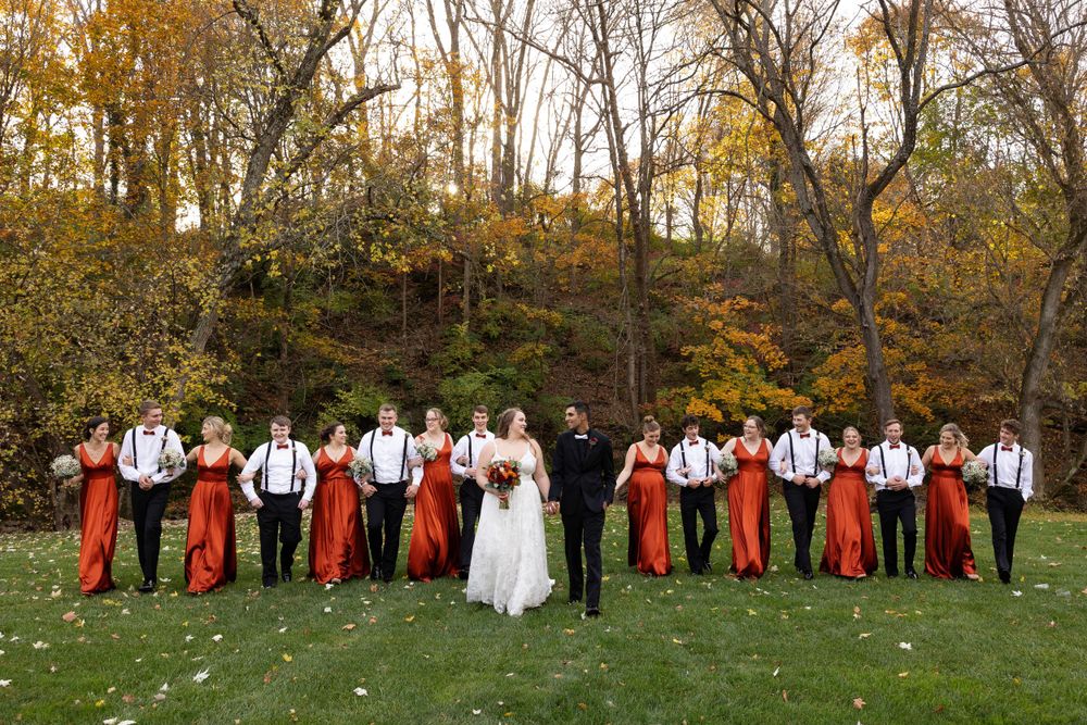 Wedding party in formal attire with red accents stand in line on grass with autumn trees behind.