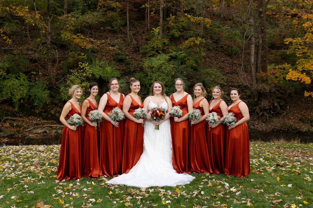 Bridal party in striking red dresses pose on autumn lawn with fall foliage backdrop.