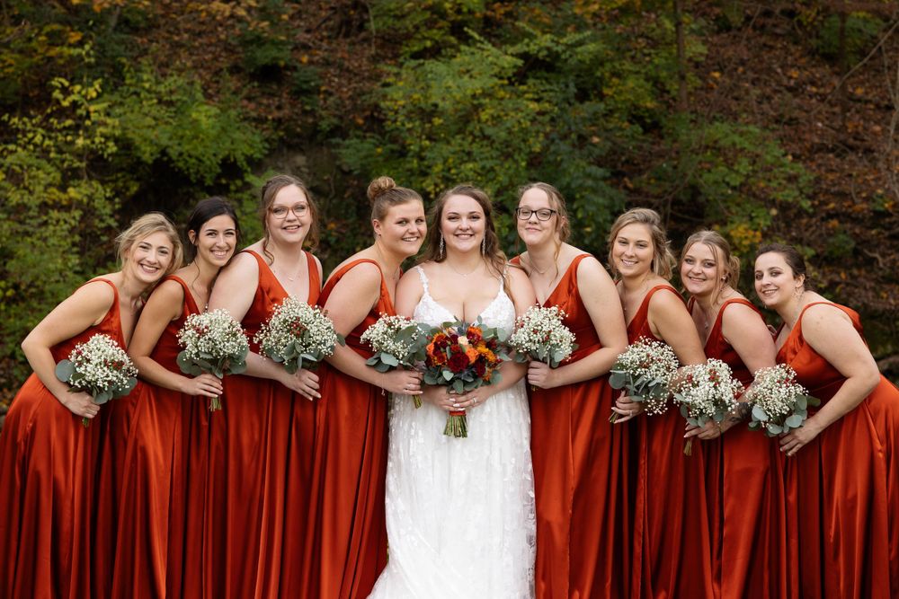 Wedding party with burgundy dresses and bouquets pose together against fall foliage.