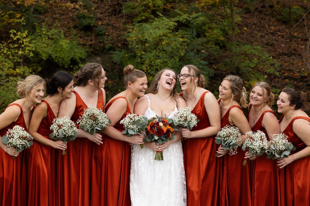 Wedding party with matching deep red dresses hold bouquets against autumn forest backdrop.