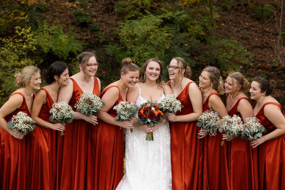Wedding party with red gowns and white bouquets share joyful moment outdoors.