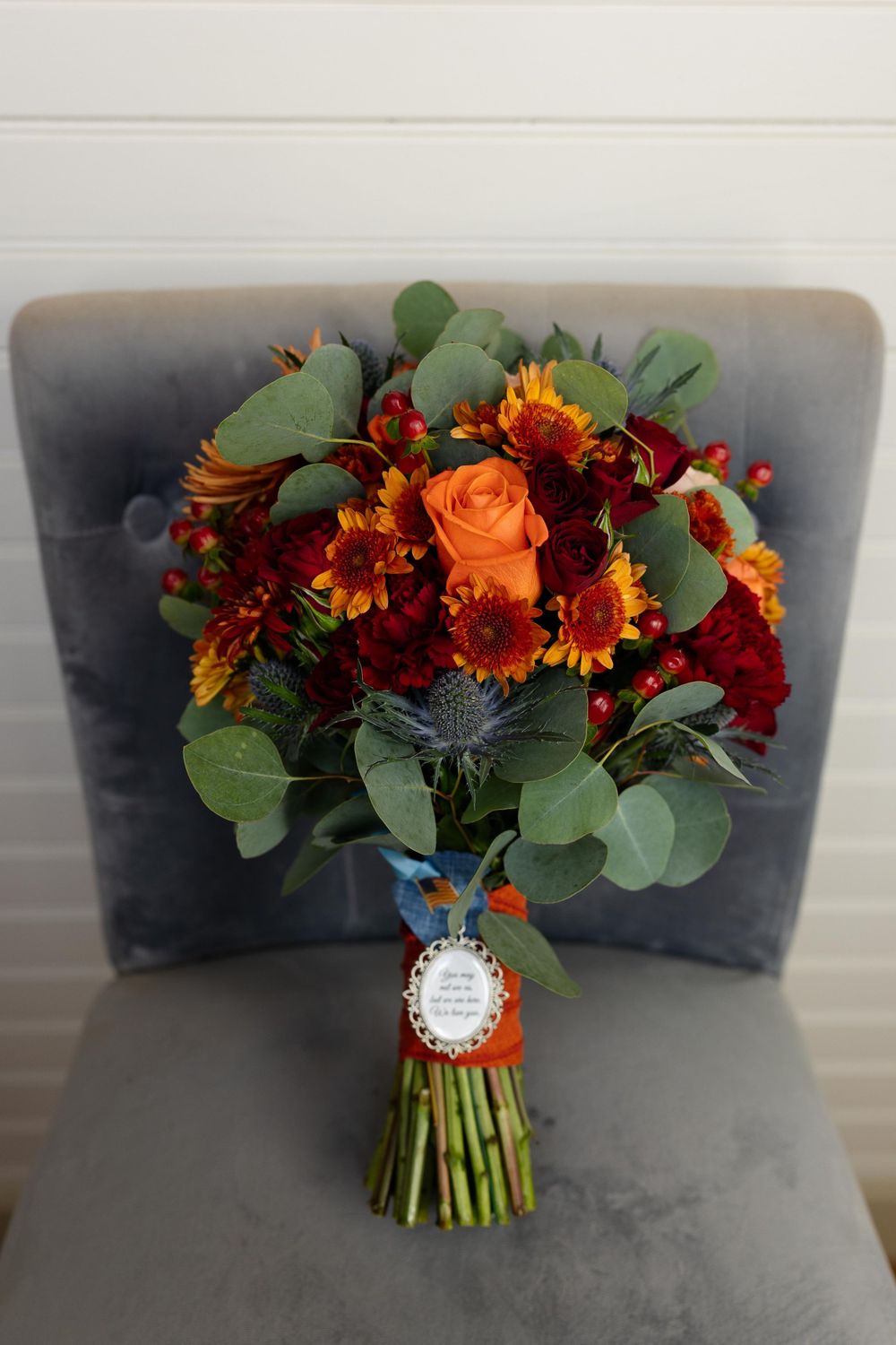 Fall wedding bouquet with orange roses, burgundy flowers and eucalyptus on grey chair.