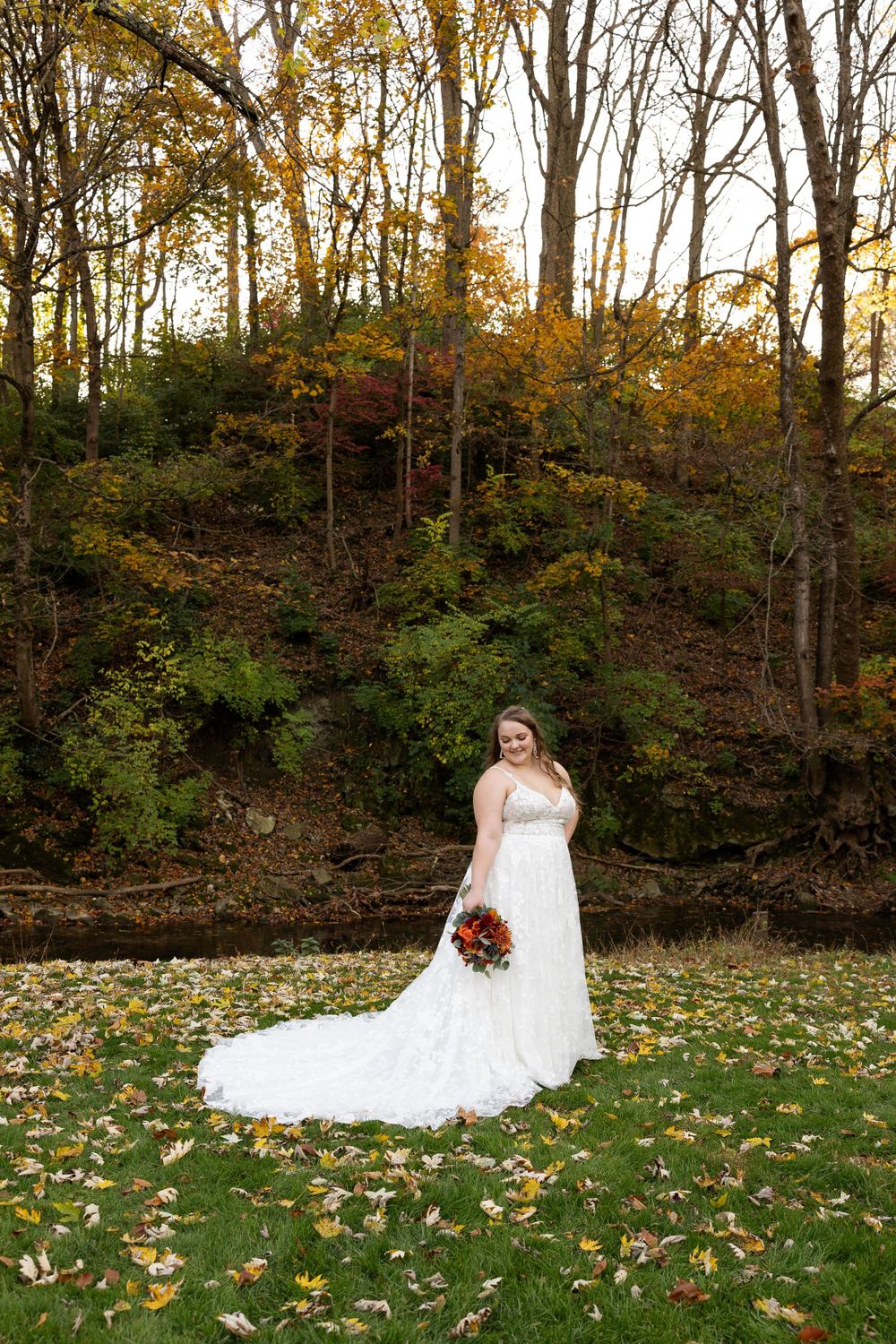 Long white wedding dress train spreads across autumn leaves with forest backdrop.