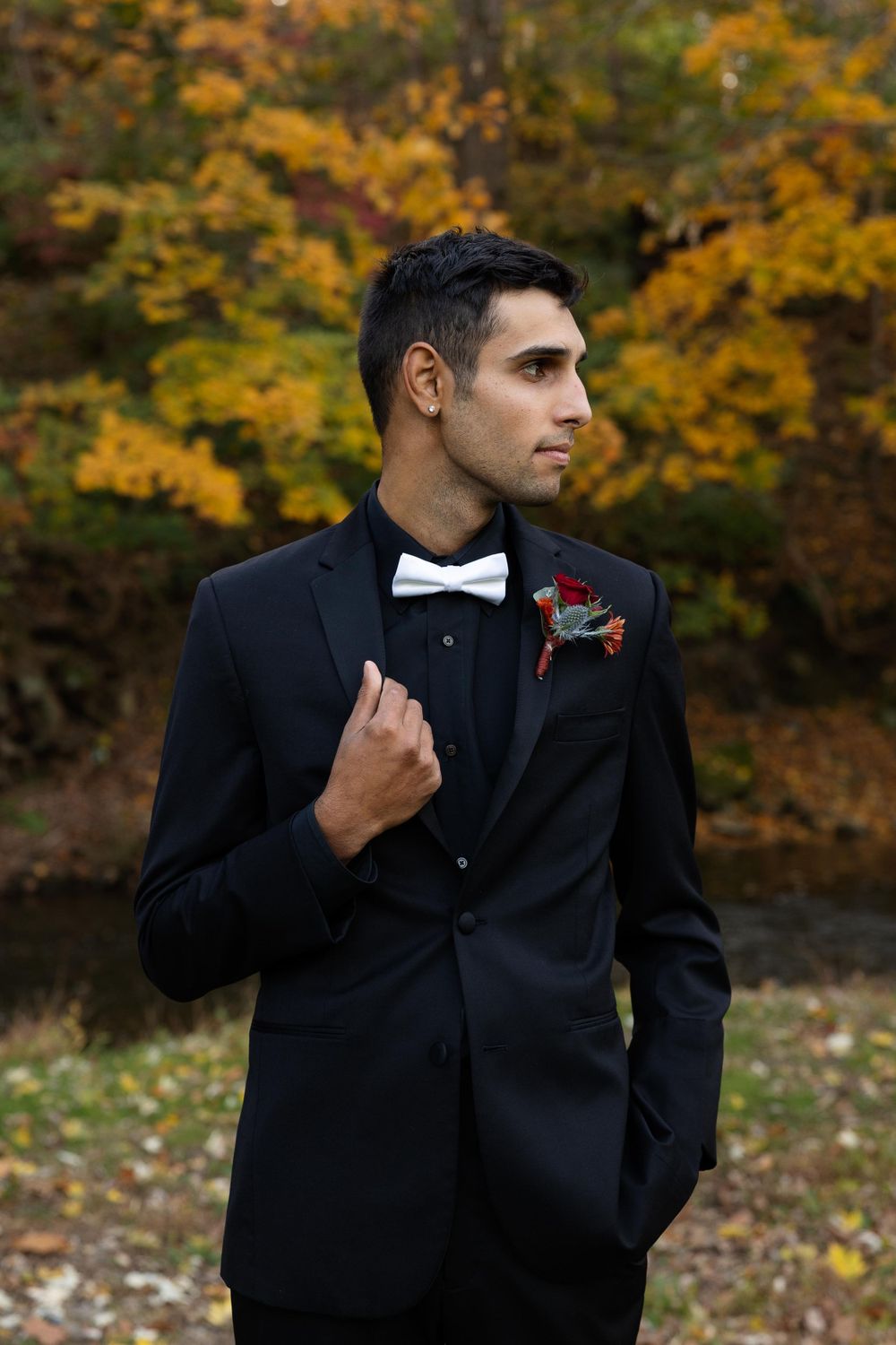 Groom in black tuxedo and bow tie with red boutonniere poses against autumn foliage background.
