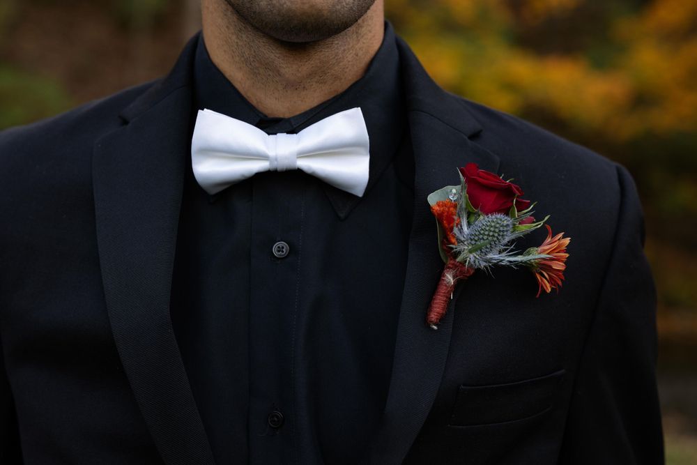 Close-up of black tuxedo with white bow tie and red rose boutonniere against fall colors.