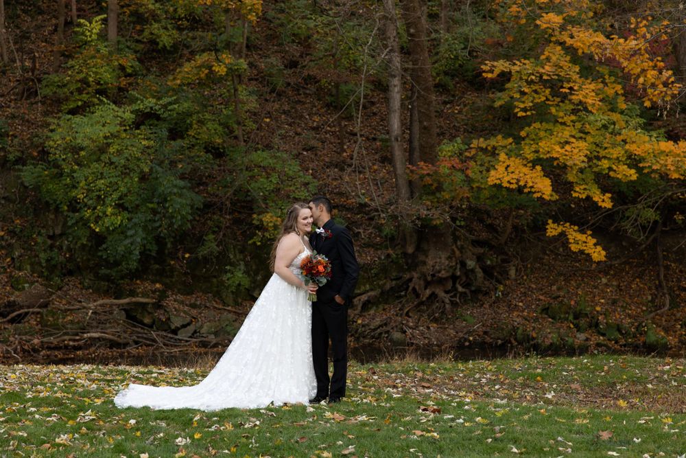 Newlyweds pose together on lawn with fall foliage and hillside in background.