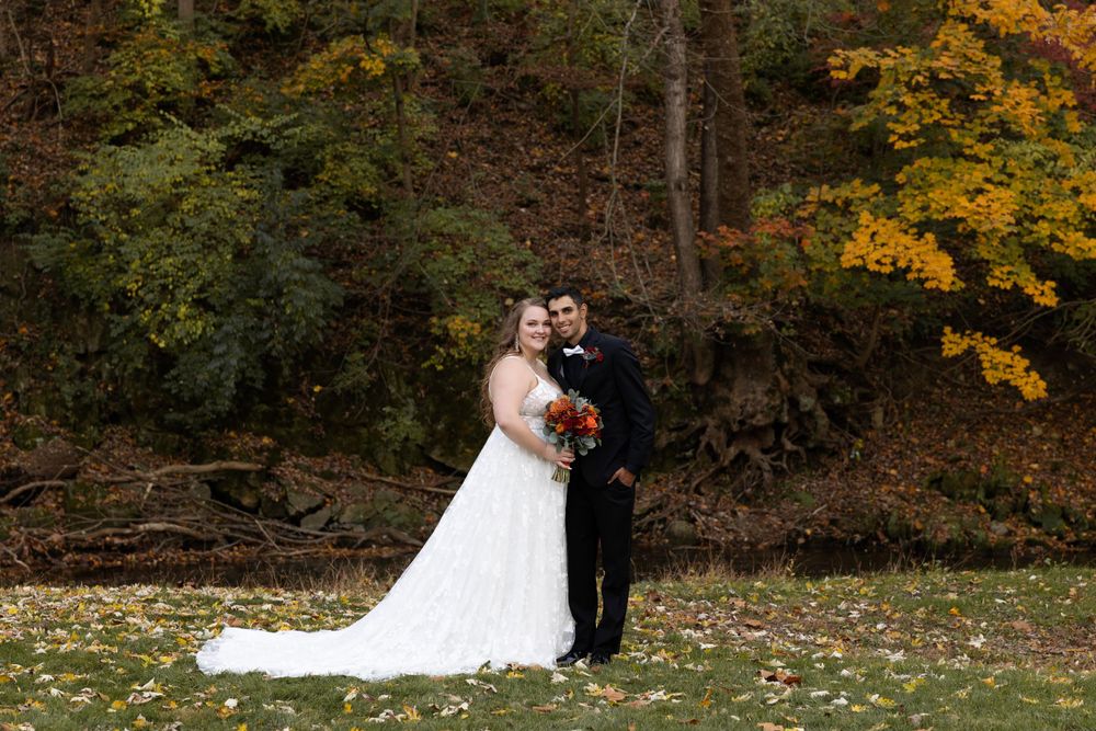 Wedding couple poses on grass with autumn trees and orange foliage behind them.