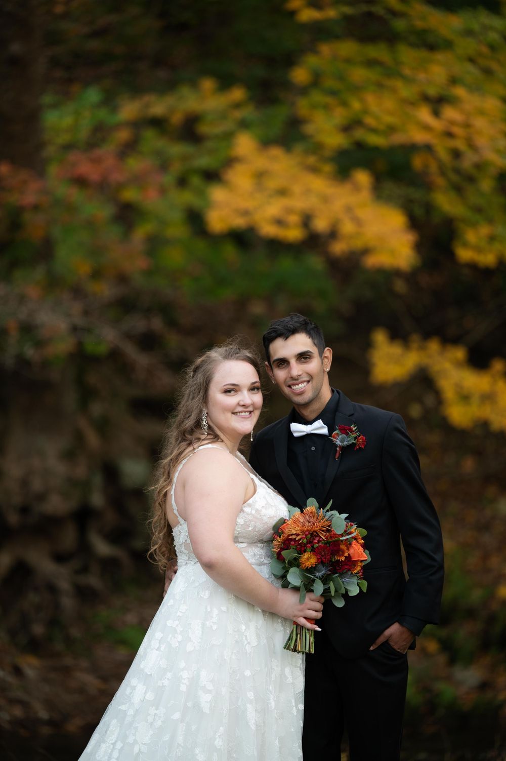 Happy newlyweds smile at camera with vibrant autumn colors in background.