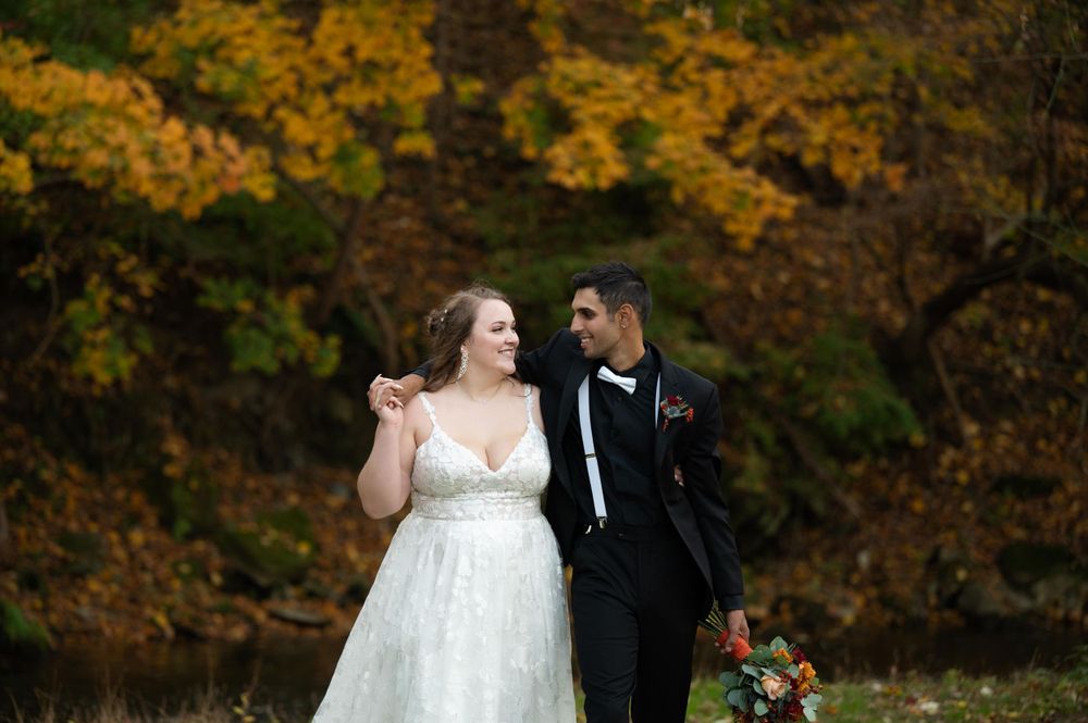 Bride in white gown and groom in tuxedo share moment against fall backdrop.
