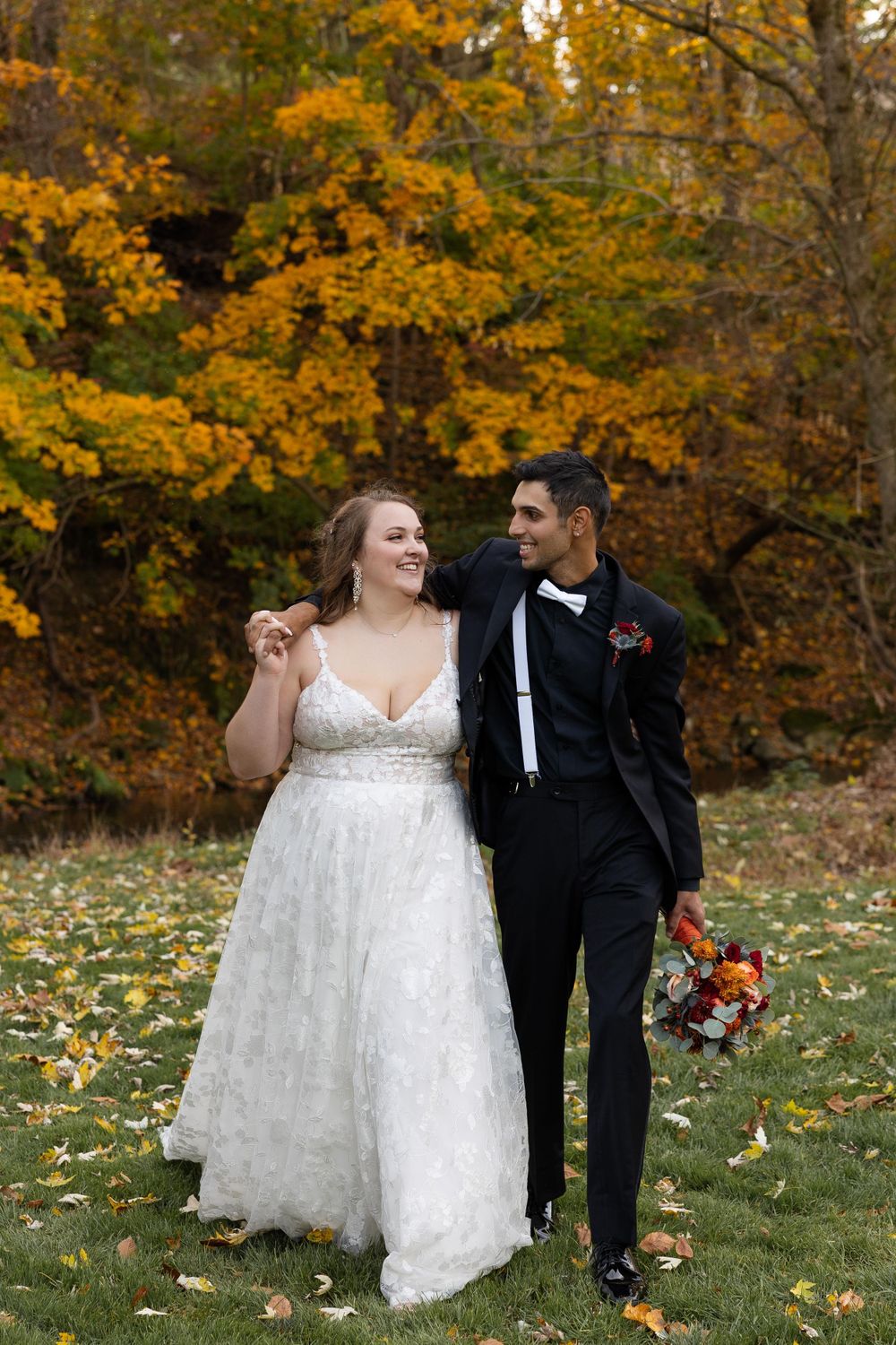 Newlyweds walking together on grass with autumn trees and fallen leaves in background.