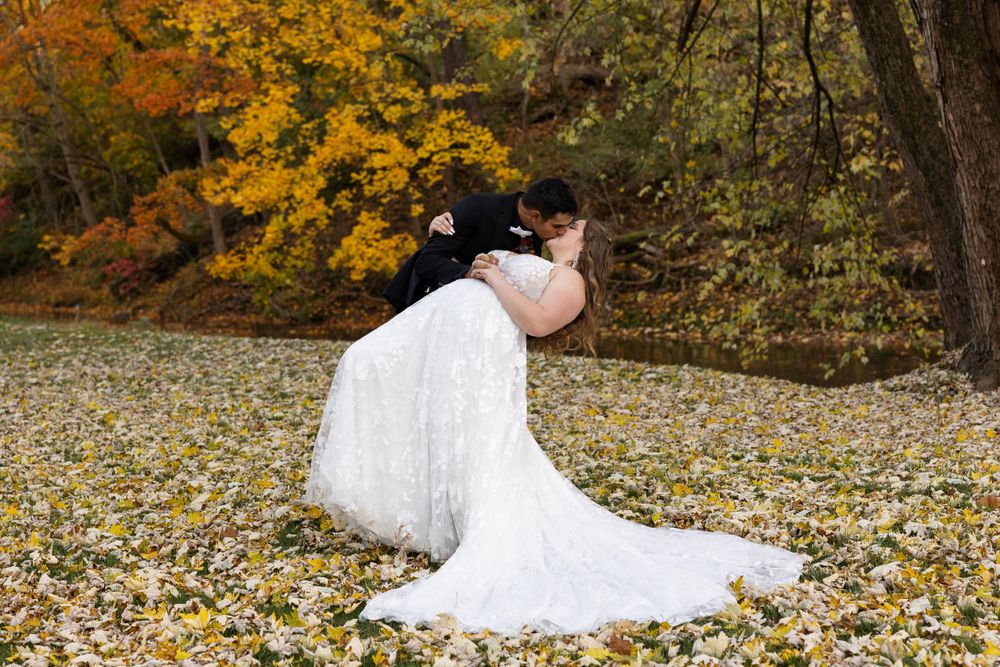 A newlywed in a white dress lying on autumn leaves while taking photos in a scenic outdoor setting.