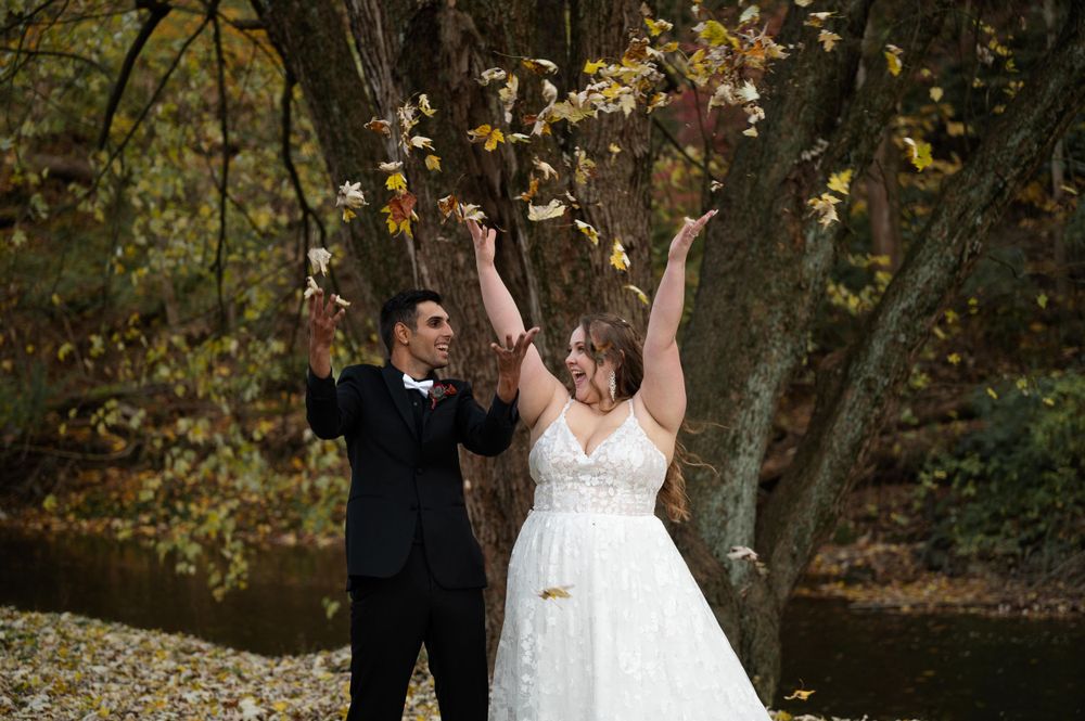 A couple celebrates joyfully outdoors surrounded by fall foliage while leaves cascade around them.