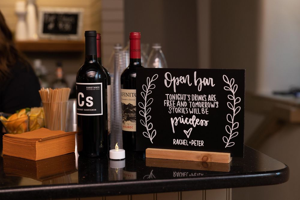 Wedding bar setup with wine bottles and decorative chalkboard sign on dark wooden surface.