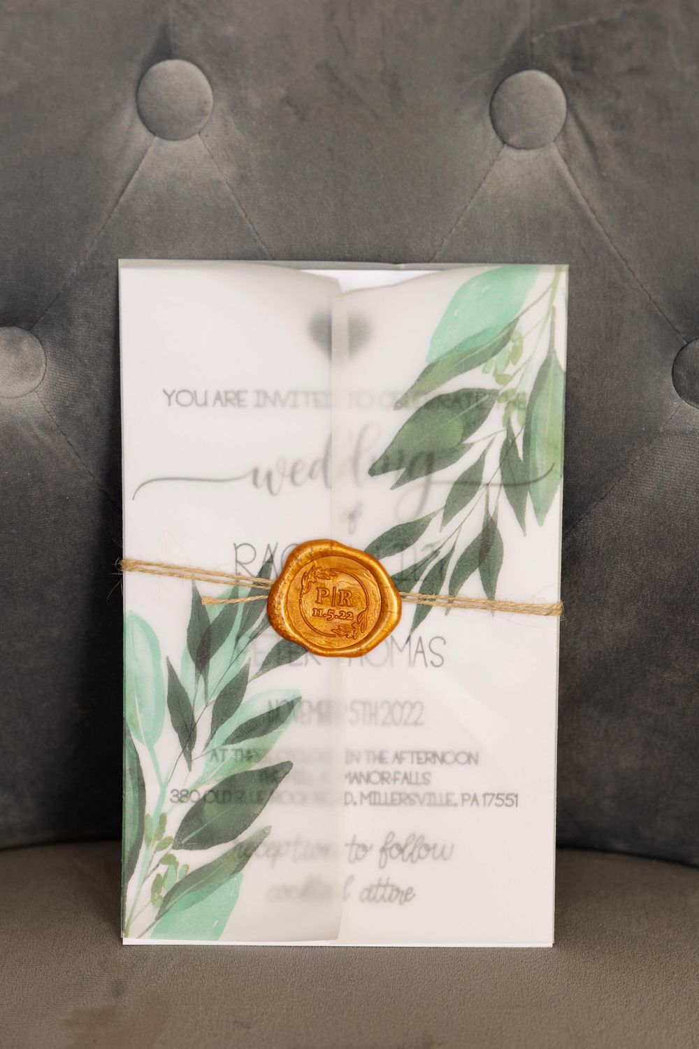 Wedding invitation with watercolor greenery and orange wax seal on grey background.
