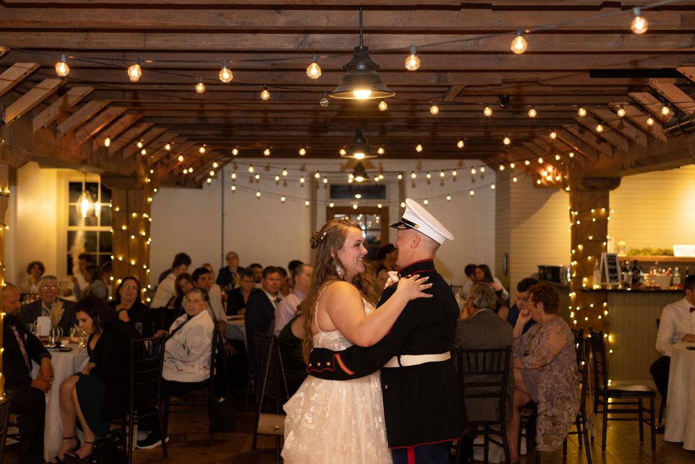 Wedding couple dancing under string lights in rustic venue with guests watching.